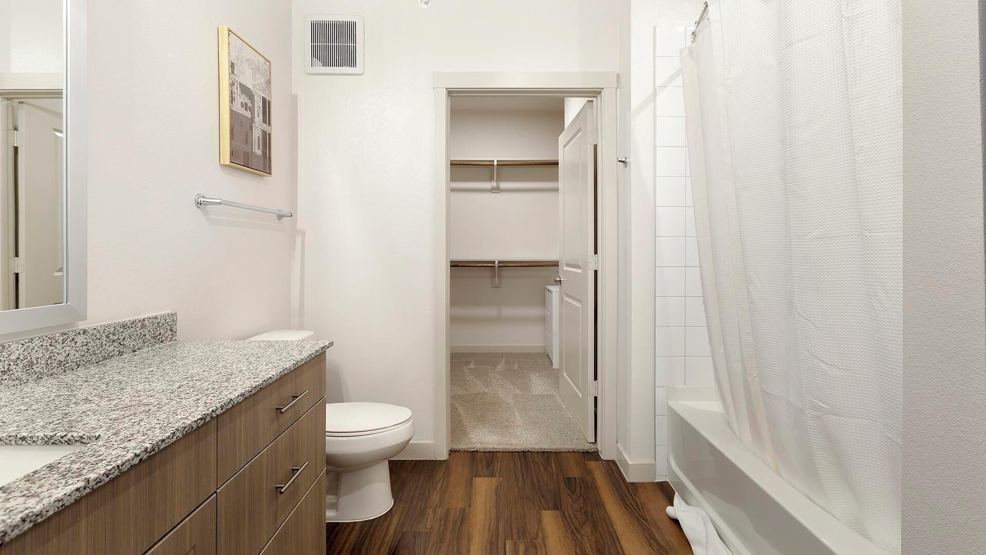 Ein Badezimmer mit einer Granit-Arbeitsplatte, Holzschränken, einer weißen Toilette, einer Dusche mit weißem Vorhang und einem begehbaren Kleiderschrank mit einem Holzboden.