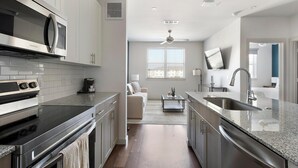 A modern kitchen with stainless steel appliances, a dining area with a sofa and coffee table, and a TV mounted on the wall.