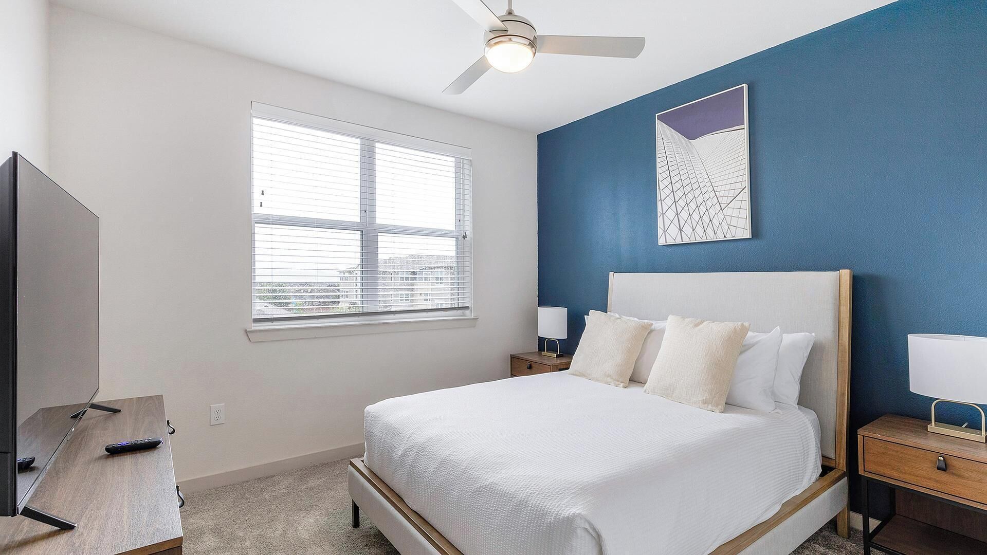 A bedroom with a bed, bedside tables, a ceiling fan, a window, and a wall-mounted artwork.
