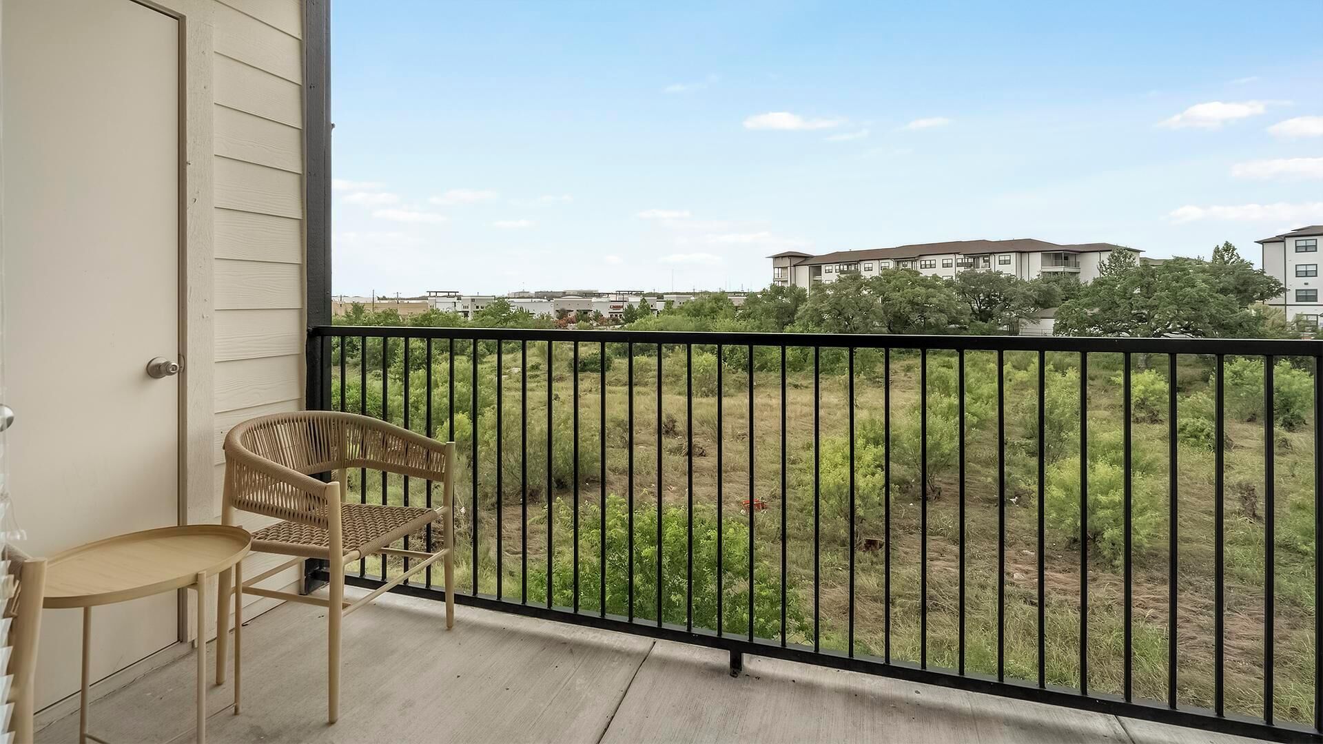 Un balcon avec une chaise en osier et une petite table, donnant sur un espace verdoyant avec des bâtiments en arrière-plan.