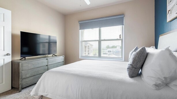 Une chambre à coucher avec un lit, un téléviseur posé sur une commode et une fenêtre avec des stores.