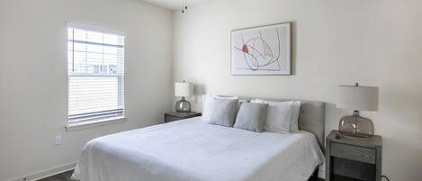 Une chambre avec un grand lit, deux tables de chevet, un ventilateur de plafond, une fenêtre avec des stores et un tableau abstrait encadré accroché au mur.