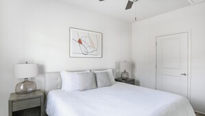 Un dormitorio con cama, dos mesitas de noche con lámparas, una puerta y un cuadro abstracto colgado en la pared.