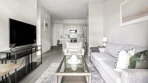 A modern living room with a flat-screen TV, a glass coffee table, a grey sofa, and a kitchen in the background. - Landing | Beautiful 1BD, Clubhouse, Gym (Round Rock)