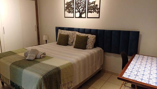 A neatly made bed with a patterned bedspread and pillows, a small table with a patterned tablecloth, and a wall with three tree decorations.