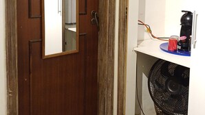 A room with a wooden door, a white wall, a small shelf with a red object, and a black fan on a countertop.