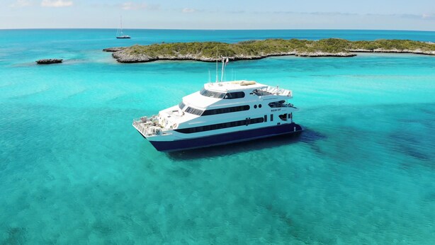 A large yacht with multiple decks, anchored in clear turquoise waters, surrounded by a small island with lush greenery.