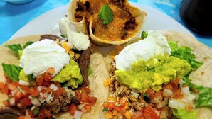 Three tacos with toppings, a bowl of sauce, and a cup of water on a table.
