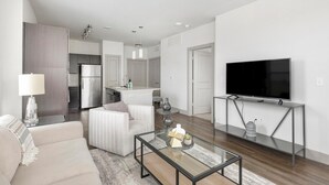 A modern living room with a glass coffee table, a sofa, a TV on a stand, and a kitchen in the background. - Landing | Chic 1BD, Yoga Studio, Pool (Haltom City)