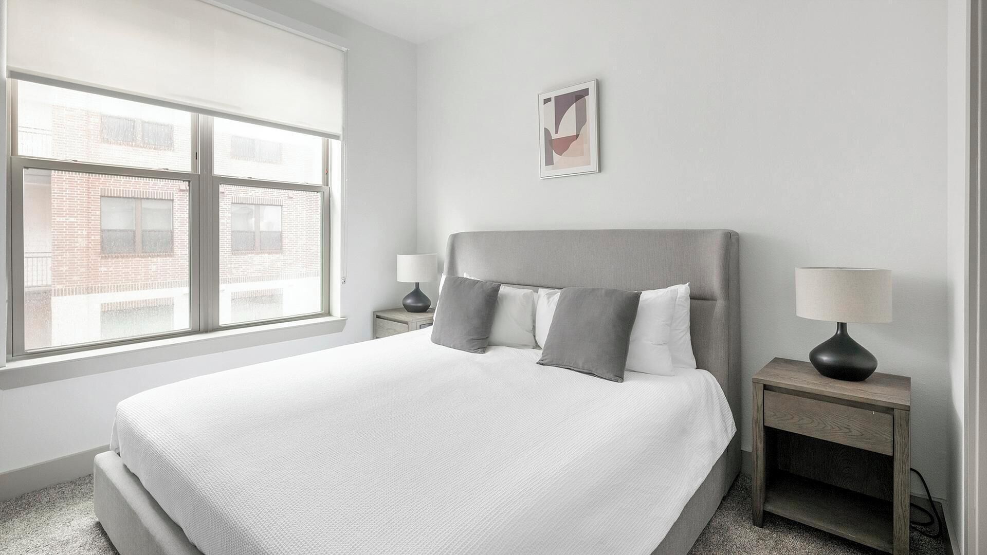 A modern hotel room with a large bed, two bedside tables, and a window with a view of a brick building.
