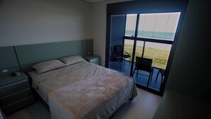 A bedroom with a large bed, a nightstand, a chair, and a view of the beach through a sliding door. - Luxury Apartment with Sea View (Matinhos)