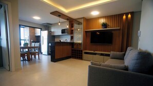 A modern living room with a grey sofa, a TV mounted on the wall, and a kitchen area with wooden cabinets and a dining table. - Luxury Apartment with Sea View (Matinhos)