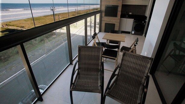 A balcony with wicker chairs, a small table, and a view of the beach and ocean. - Luxury Apartment with Sea View (Matinhos)