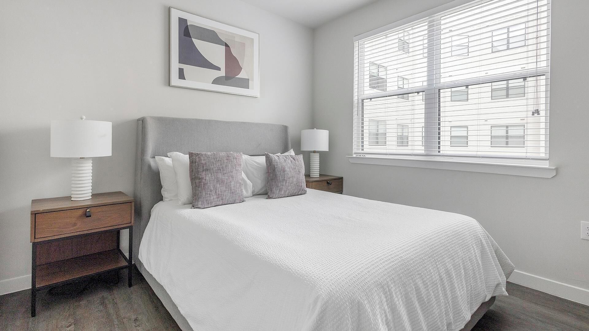 A bedroom with a large bed, two bedside tables, a window with blinds, and a framed abstract painting on the wall.