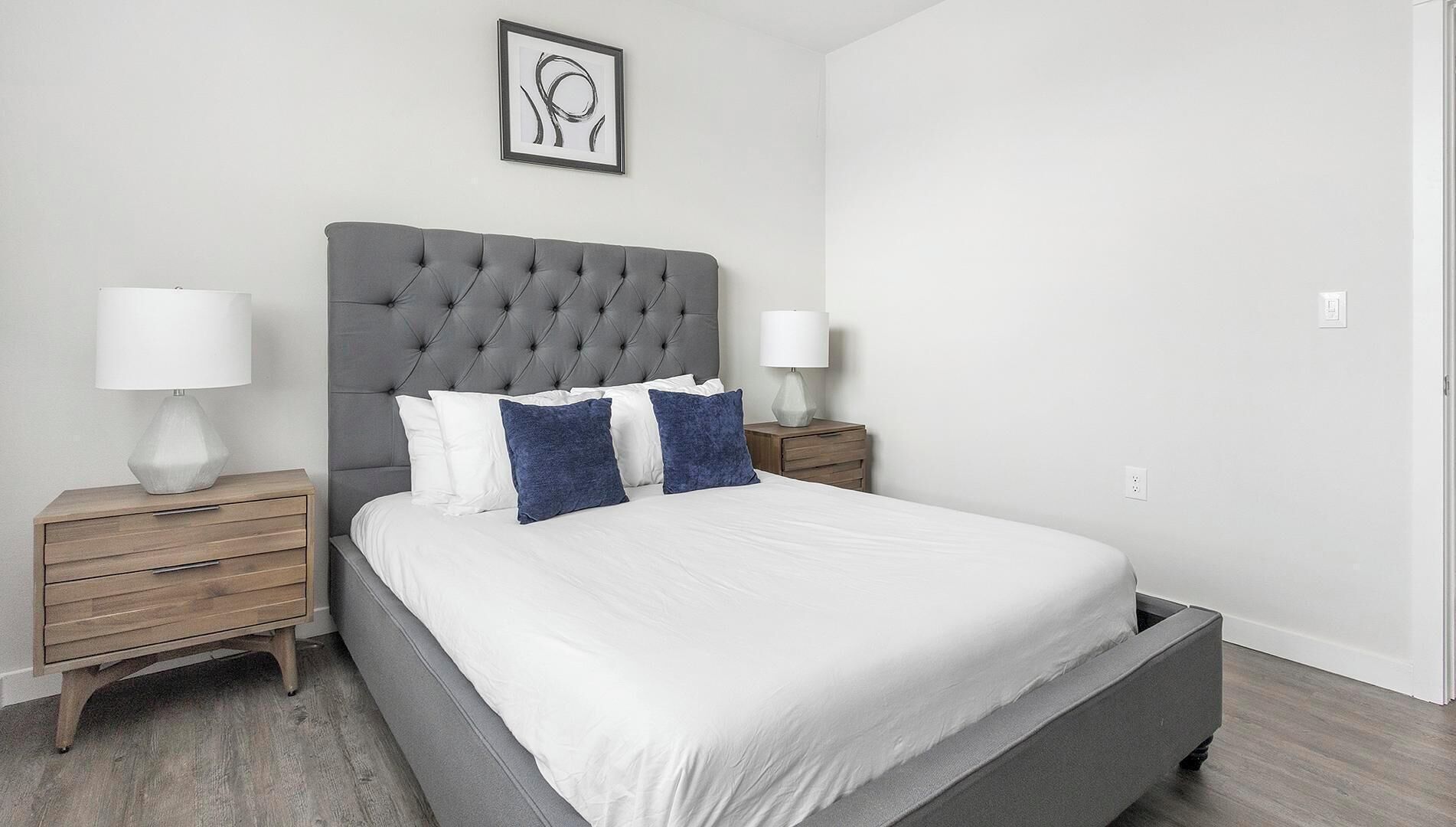 A modern bedroom with a grey tufted headboard, white bedding, and two bedside tables with lamps.