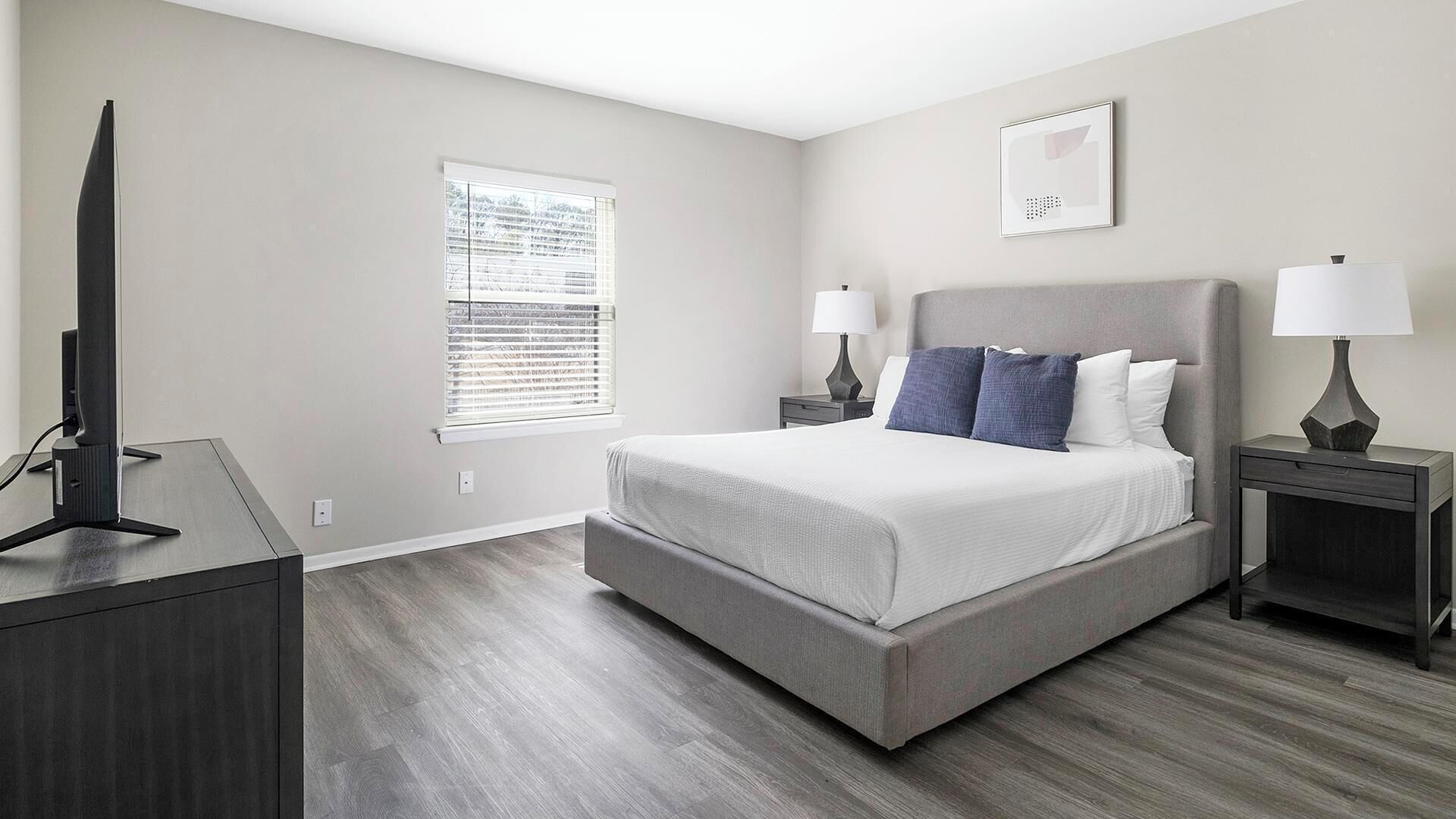 A modern bedroom with a large bed, two bedside tables, a TV on a stand, and a window with blinds.