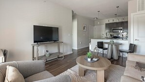 Ein modernes Wohnzimmer mit Flachbildfernseher, Couchtisch und Sofa. Dazu gehört eine Küchenzeile mit Mikrowelle sowie ein Esstisch mit Stühlen.