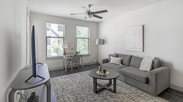 A modern living room with a grey sofa, a round coffee table, a TV on a stand, a ceiling fan, and two windows with blinds.