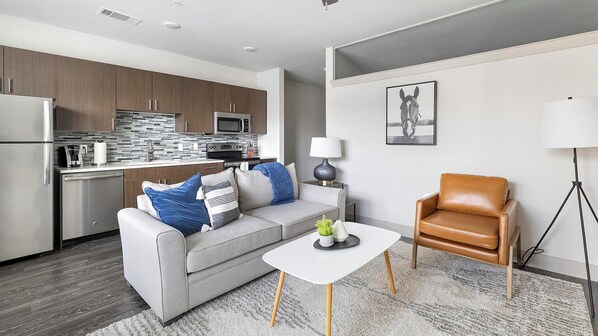 A modern living room with a grey sofa, a coffee table, a brown leather chair, and a kitchen in the background. - Landing | Incredible 1BD, Pool, Gym (Dallas)