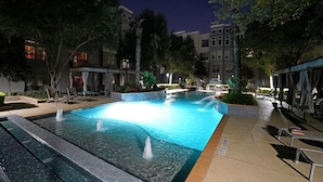 A nighttime swimming pool area with multiple water features, lounge chairs, and a well-lit environment.