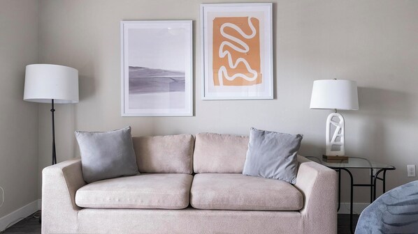 Un sofá color beige con dos almohadones, una lámpara de pie y dos cuadros en la pared.