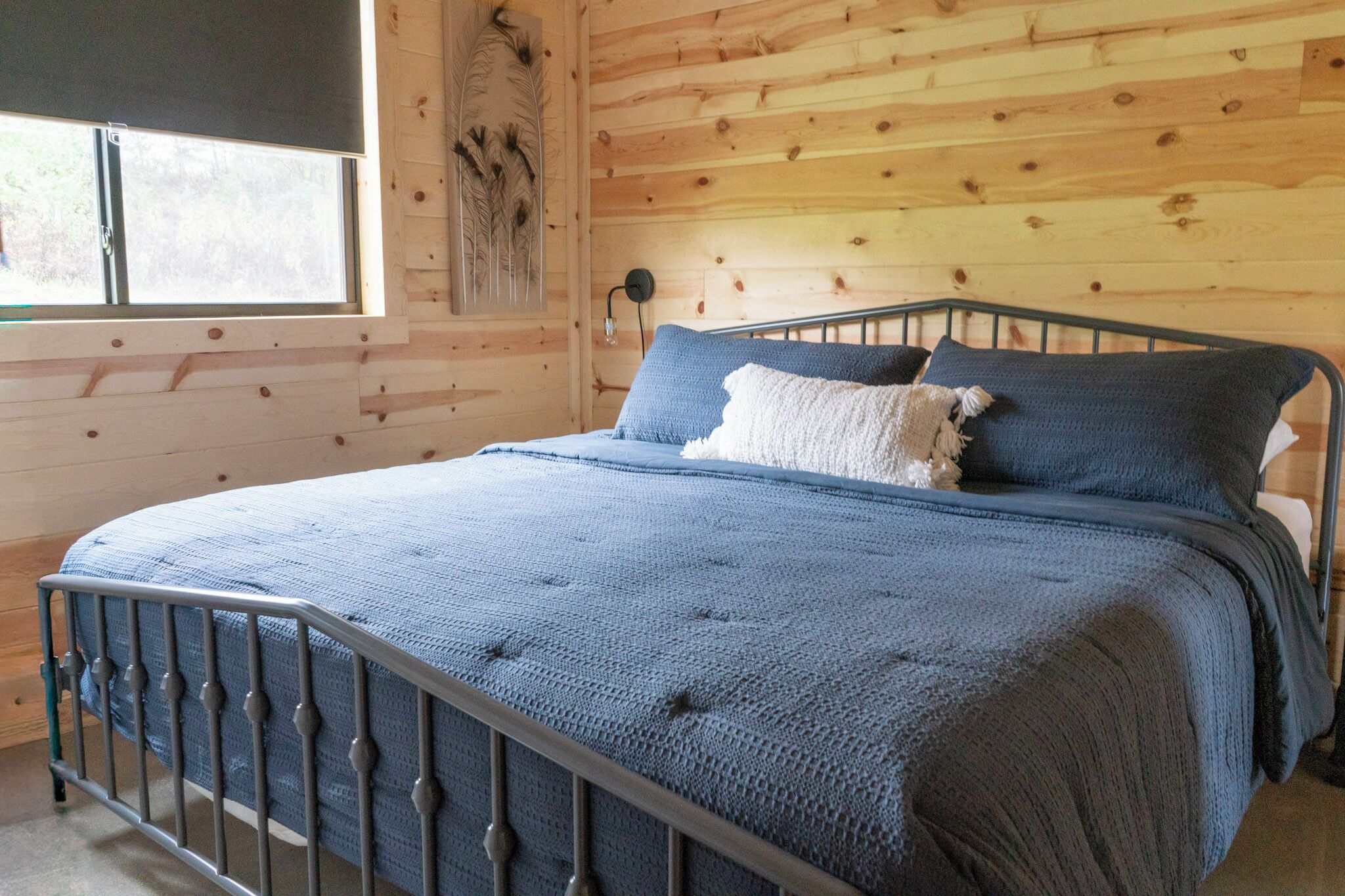 Una cama con estructura metálica, una manta azul y dos almohadas en una habitación con paredes de madera y una ventana.