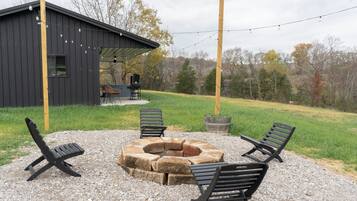 A covered outdoor seating area with a fire pit, surrounded by benches and a rustic building in the background.