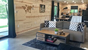 A modern living room with a wooden ceiling, a sofa, a coffee table, and a kitchen area in the background. - The Barrel Proof Bourbon Trail Cottage Private Hot Tub Firepit Lawrenceburg Ky. (Lawrenceburg)