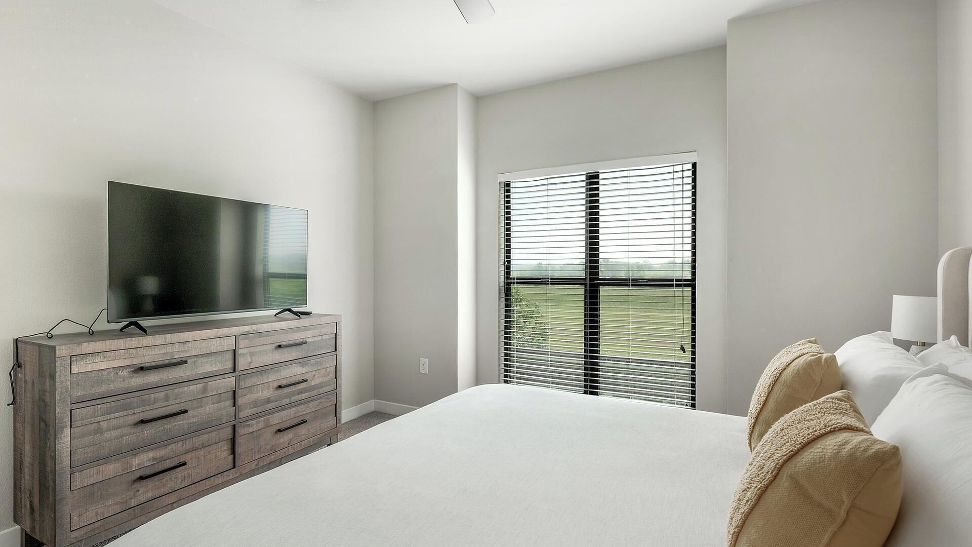 A bedroom with a large bed, a wooden dresser, a flat-screen TV, and a window with blinds.