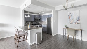 A modern kitchen with a central island, bar stools, stainless steel appliances, and a small dining table with a chair. - Landing | Stylish 1BD, Game Room, Pool (Haltom City)