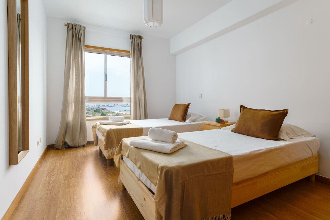 A hotel room with two beds, wooden flooring, a large window with curtains, and a view of the outdoors.