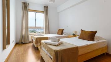A hotel room with two beds, wooden flooring, a large window with curtains, and a view of the outdoors.