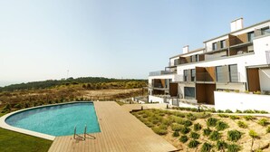 A modern two-story building with a swimming pool, a wooden deck, and a view of the beach.