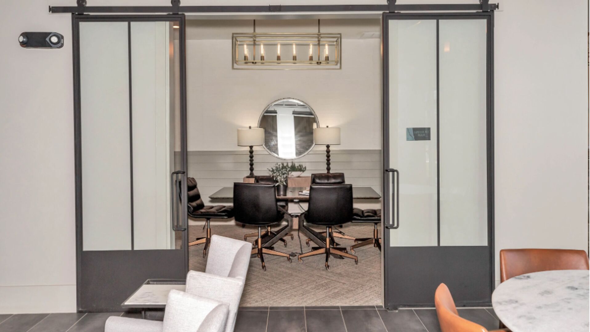 A modern dining area with a glass partition, a round mirror, two lamps, and a table with chairs.