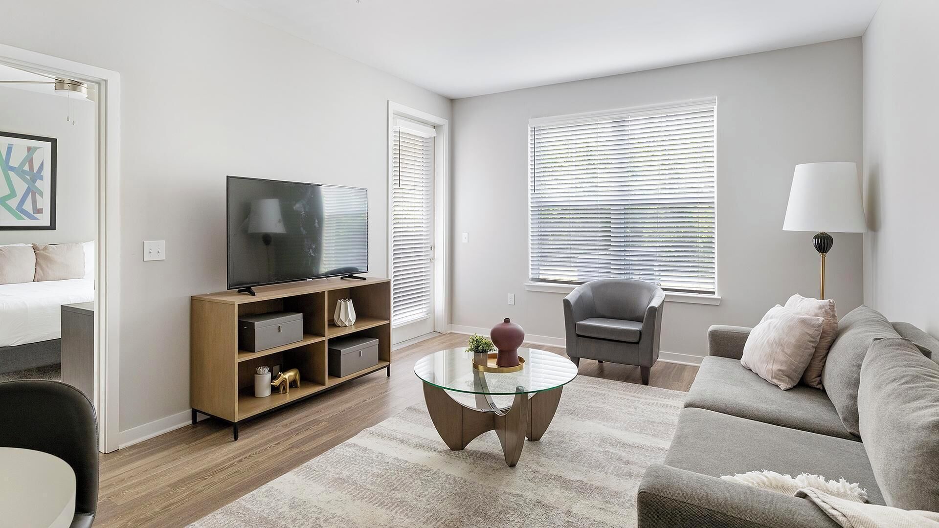 A modern living room with a flat-screen TV, a glass coffee table, a grey sofa, and a grey armchair.