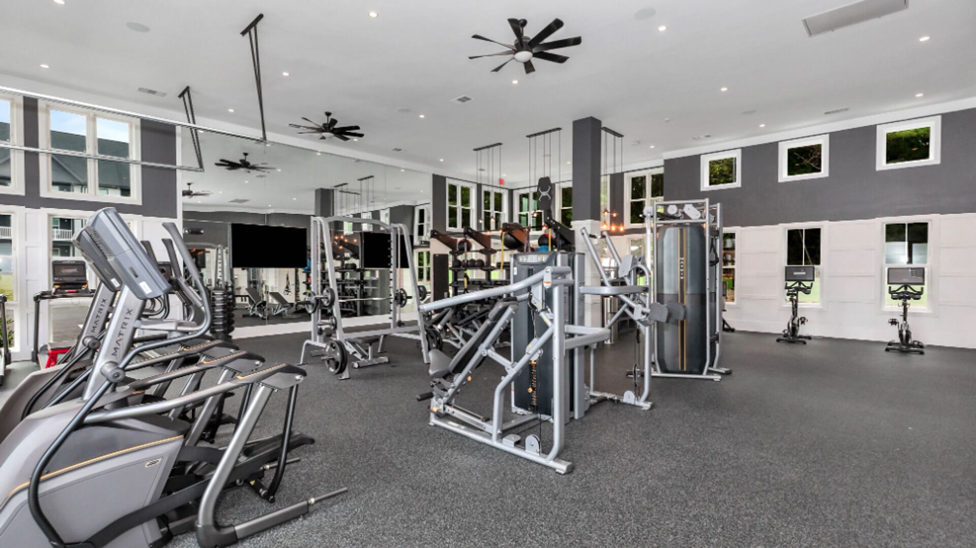 Un gimnasio moderno con varios equipos de ejercicio, ventanas amplias y un ventilador de techo.