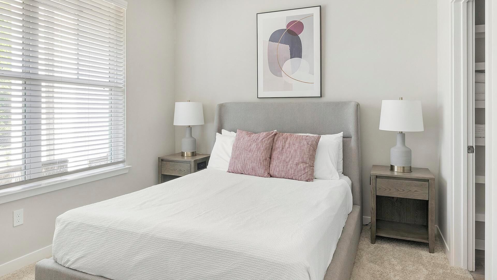 Quarto bem organizado com uma cama, duas mesas de cabeceira com luminárias, uma janela com persianas e um quadro abstrato pendurado na parede.