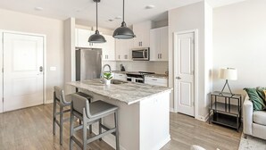 Une cuisine moderne avec des meubles blancs, un îlot central doté de tabourets de bar, des appareils en acier inoxydable et un éclairage suspendu.