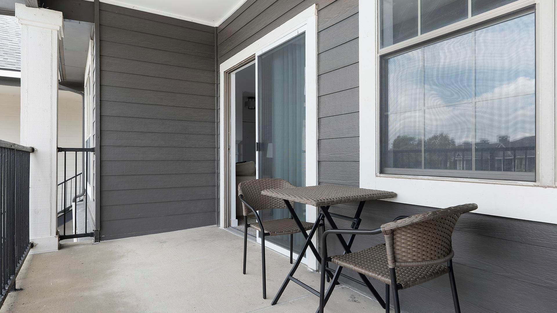 A balcony with two wicker chairs and a small table, a sliding glass door, and a large window.