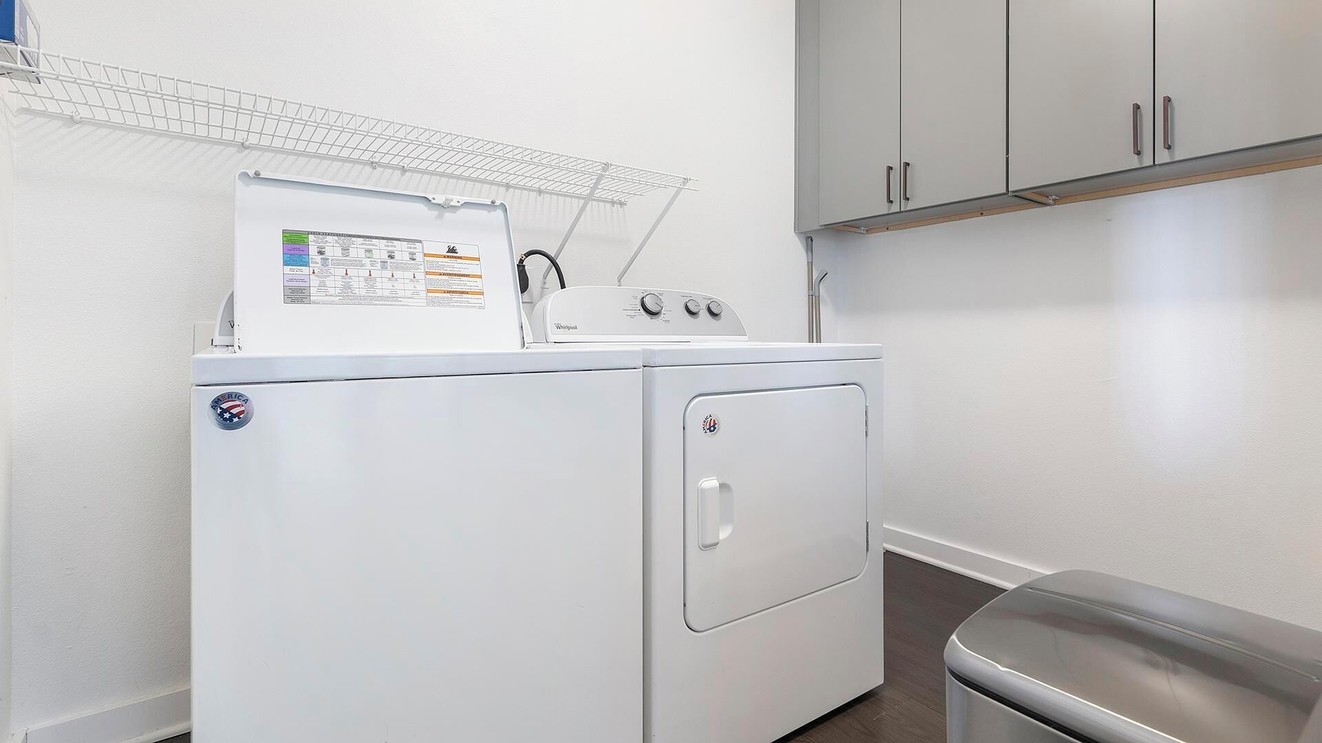 A compact laundry room with a front-loading washer and dryer, a drying rack above the washer, and a trash bin to the right.