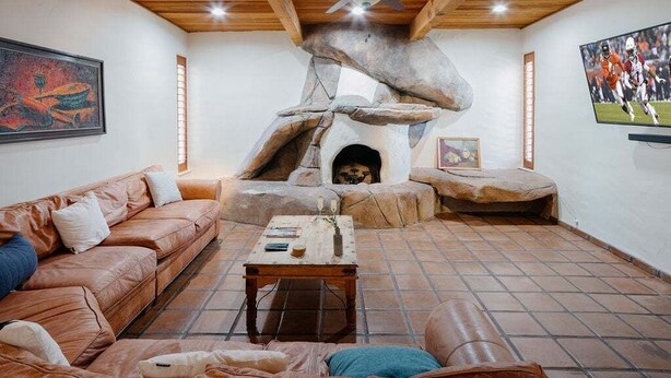 A cozy living room with a stone fireplace, leather sofas, and a ceiling fan. - Bed Rock Desert -pool Heat Available - Spring Break (Cave Creek)