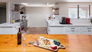 A modern kitchen with white cabinetry, a wooden dining table with a charcuterie board, and a view of the outdoors through large windows. - Bed Rock Desert -pool Heat Available - Spring Break (Cave Creek)