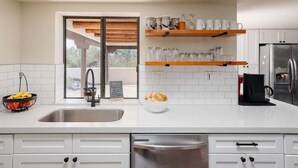 A modern kitchen with white cabinets, a stainless steel sink, and a built-in dishwasher. There are wooden shelves above the sink holding glassware and a bowl of bread. - Bed Rock Desert -pool Heat Available - Spring Break (Cave Creek)