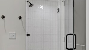 A modern bathroom with white tiled walls, a glass shower enclosure, and a beige rug. - Euphony-designer Furnishings-5 Mins to Broadway (Nashville)