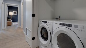 Et vaskerum med to frontbetjente Whirlpool vaskemaskiner, en dør til et opholdsområde med sofa og lampe, samt en gang med lyse gulve.
