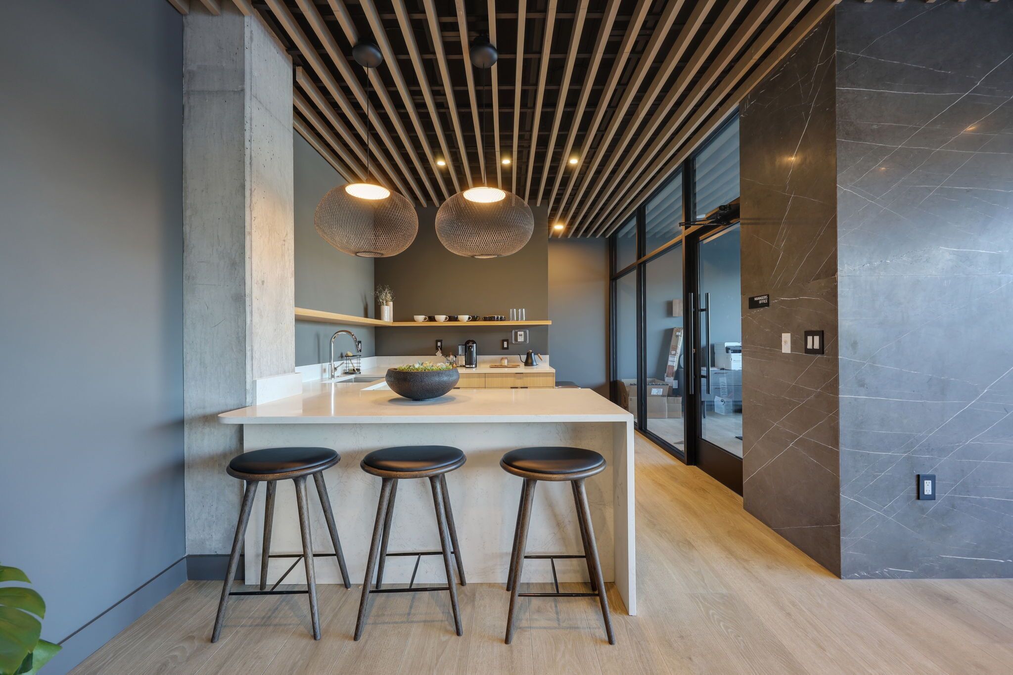 Cocina moderna con una isla central, taburetes de bar, iluminación colgante y vistas al área de oficina.