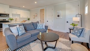 Una sala moderna con un sofá gris, una mesa de centro redonda y un sillón blanco. Hay una cocina con armarios blancos y un microondas.