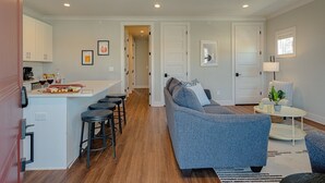 A modern kitchen with white cabinets, a dining area with bar stools, a grey sofa, and a small round table with a plant. - Modern Melrose-sleeps 8- Mins to Downtown Broadway (Nashville)