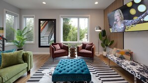 A modern living room with a green sofa, two brown armchairs, a blue ottoman, a black and white striped rug, and a large TV on the wall. - Vivid Vocals - 4 Mins to Broadway - Rooftop Patio (Nashville)