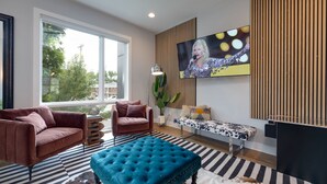 A modern living room with a large window, a television mounted on the wall, and a mix of furniture including a sofa, armchair, and ottoman. - Vivid Vocals - 4 Mins to Broadway - Rooftop Patio (Nashville)